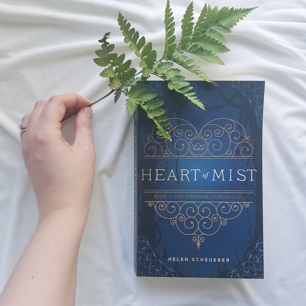 A hand holds a fresh fern leaf over the blue cover of Heart of Mist